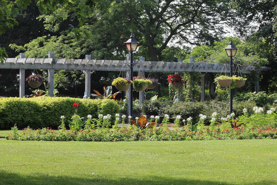 A well-maintained garden featuring a lush green lawn in the foreground, bordered by a variety of colorful flowers including red, white, and pink blooms. Behind the flower bed, there are neatly trimmed