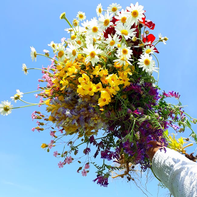 A vibrant bouquet of fresh flowers held up against a clear blue sky, featuring white daisies with yellow centers, bright yellow primulas, purple statice, and small pink and blue blooms. The arrangemen