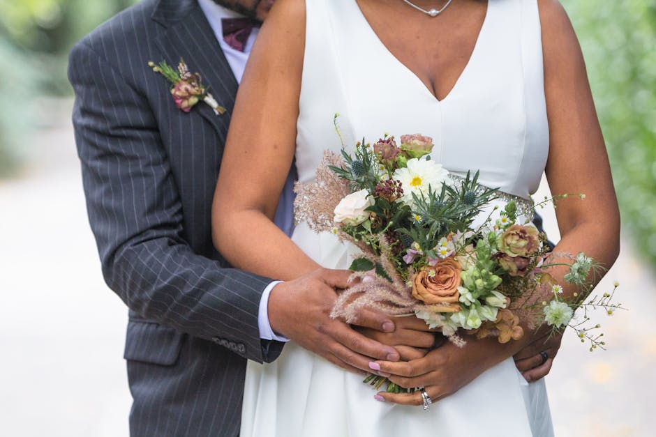 Close-up of a bride and groom holding a floral bouquet during a wedding, with the bride wearing a white dress and the groom in a dark pinstriped suit with a boutonnière. The bouquet features a mix of