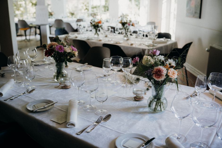 A close-up view of a wedding floral arrangement placed on a white tablecloth in an elegant event setting. The bouquet in a clear glass vase features a mix of fresh flowers including pink and white car