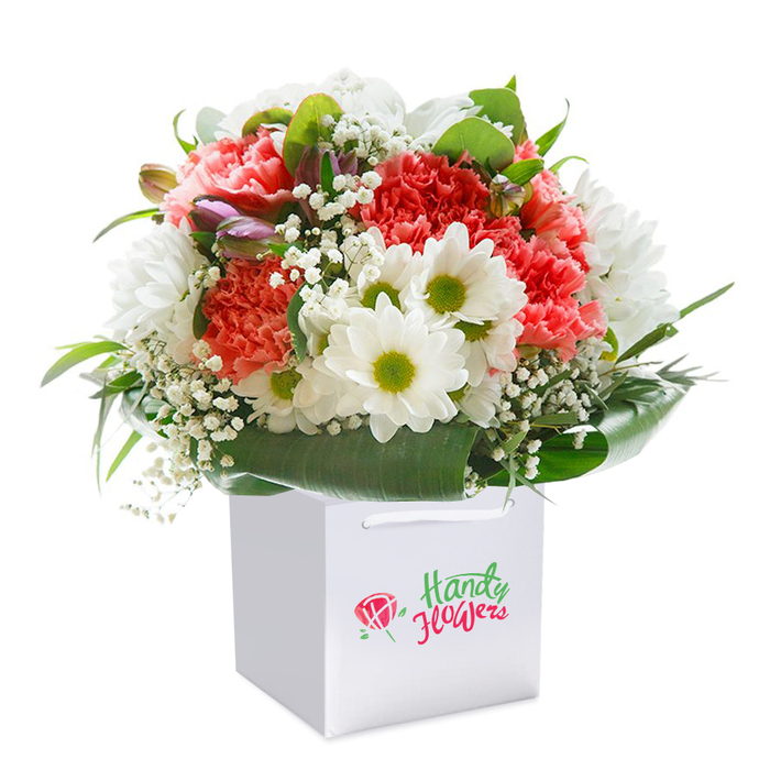 Presented upright in an immaculately white square gift bag, this floral arrangement bursts with freshness and colour, the hallmark of Dartmouth Park Florist's commitment to quality and style. The core blooms include lush coral-pink and peach carnations, instantly recognizable by their abundant ruffled petals, mingling harmoniously with clusters of bright white daisies each tipped with a sunny lime-green centre, reminiscent of the cheerful floral displays often seen outside homes on Chester Road. Wisps of white baby's breath are artfully tucked throughout, providing both lightness and a subtle structure, while several closed buds tinged with purple peek out from the left side, suggesting the promise of new growth. Deep green, broad leaves gracefully arc around the base, encasing the arrangement in a soft yet striking natural border that enhances its visual impact. The bouquet's overall form is pleasingly rounded-carefully crafted to convey both exuberance and refinement. Set against a clean, unobtrusive white backdrop, the flowers and packaging alike exude an aura of sophistication and freshness. Designed with the community of Dartmouth Park in mind, and suitable for delivery to homes from Swain's Lane to Gospel Oak, this bouquet is perfect for marking life's celebrations-be they birthdays, anniversaries, or spontaneous gestures of gratitude. Its elegant yet joyful presence makes it especially fitting for those who cherish the vibrant yet gentle spirit of North London's residential neighbourhoods.