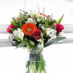 Nestled in a crystal-clear glass vase, this evocative floral creation is a testament to balanced colour, expert design, and emotional warmth-a fitting tribute to the gentle vibrancy found throughout Dartmouth Park. Luminous peach and orange gerberas ring the centre, their soft petals fanning out in a halo of light and warmth, like spring sun upon the pathways of Waterlow Park. Rich, fuchsia roses cluster close, their deep tones adding depth and a hint of romantic intrigue, while fine sprays of white-alstroemeria, perhaps tiny spray roses-brighten pockets within the arrangement. Slender aspidistra leaves sweep around the bouquet's base, forming a protective cradle that mirrors the greenery of nearby gardens. Between them, pointed leaves and trailing tendrils of foliage weave subtle patterns, contributing both volume and background contrast. The entire bouquet is invigorated by light, streaming in like morning sun through a Dartmouth Park bay window, drawing out each colour and texture without overwhelming the individual blooms. Inhale and imagine a fragrant blend-sweet rose, lily, fresh-cut greenery-wafting through the room, evoking the freshness of a newly opened florist on York Rise. Ideal for uplifting a living space, welcoming friends to brunch, or expressing gratitude, this bouquet is a reflection of optimism and considered care, as if each stem were selected to brighten life in Dartmouth Park.